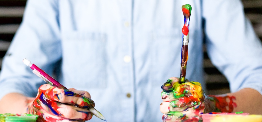 Man in a blue button down shirt with paint on his hands and holding a paintbrush